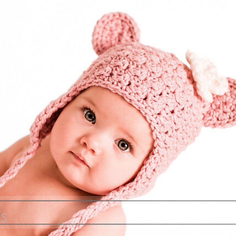 May include: A baby wearing a pink crocheted hat with bear ears and a white flower accent. The hat has long ties that can be tied under the chin.