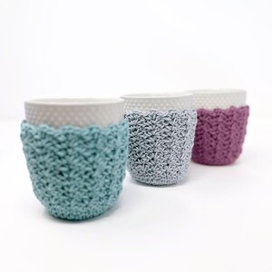 May include: Three ceramic cups with textured rims, each adorned with a crocheted cozy. The cozies are in shades of teal, gray, and purple. The cups are arranged in a row against a white background.