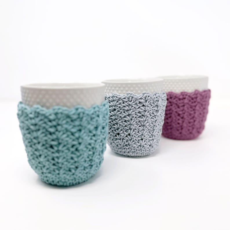 May include: Three ceramic cups with textured rims, each adorned with a crocheted cozy. The cozies are in shades of teal, gray, and purple. The cups are arranged in a row against a white background.