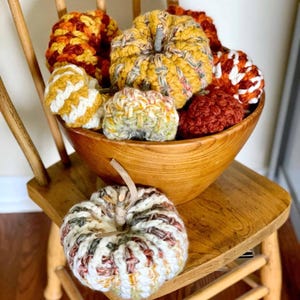 May include: A collection of handmade crochet pumpkins in various sizes and autumn colors including orange, yellow, white, and brown. Several pumpkins are contained within a large wooden bowl, while one pumpkin rests on the wooden chair seat below.