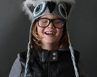 May include: A gray and white crocheted owl hat featuring large circular eyes with blue, white, and black yarn details, pointed ear flaps with fuzzy trim, and long braided tassels hanging down from each side of the head covering.