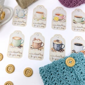 May include: A collection of decorative tags with watercolor illustrations of coffee and tea cups. Each tag has the text "a crocheted hug for your mug". The tags are arranged with a blue crocheted mug cozy and wooden buttons.