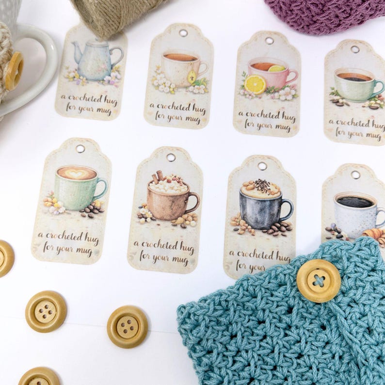 May include: A collection of decorative tags with watercolor illustrations of coffee and tea cups. Each tag has the text "a crocheted hug for your mug". The tags are arranged with a blue crocheted mug cozy and wooden buttons.