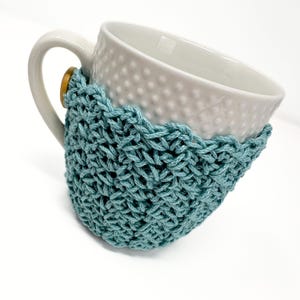 May include: A white ceramic mug with a textured surface is partially covered by a teal crocheted cozy. The cozy has a scalloped edge and is secured with a gold button. The mug's handle is visible on the left side.