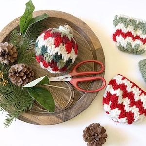 May include: A handmade Christmas ornament with a red, green, and white crocheted pattern sits on a wooden tray with pinecones, greenery, and red-handled scissors. Two additional crocheted items are also visible.
