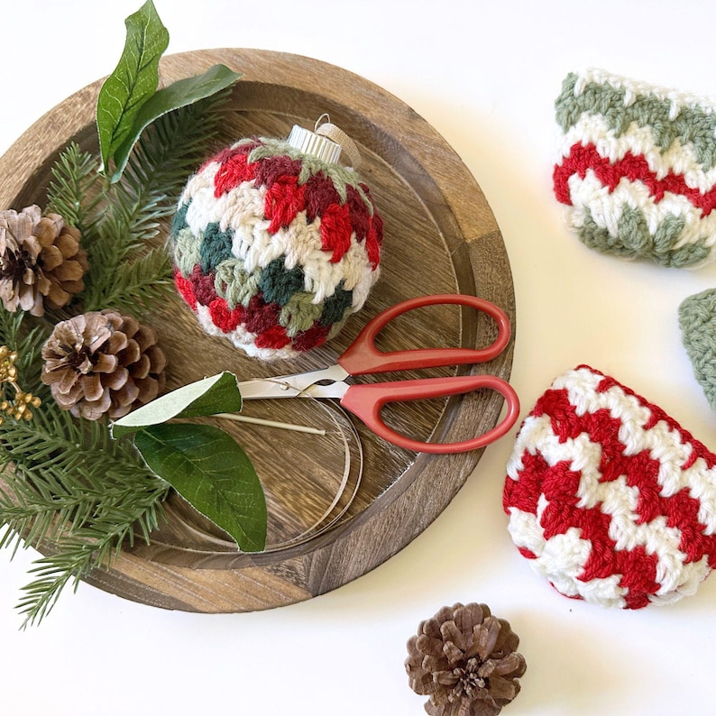 May include: A handmade Christmas ornament with a red, green, and white crocheted pattern sits on a wooden tray with pinecones, greenery, and red-handled scissors. Two additional crocheted items are also visible.