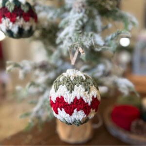 May include: Crocheted Christmas ornaments in red, white, and green. The handmade ornaments are hanging on a snow-covered Christmas tree. The ornaments have a textured, zig-zag pattern.