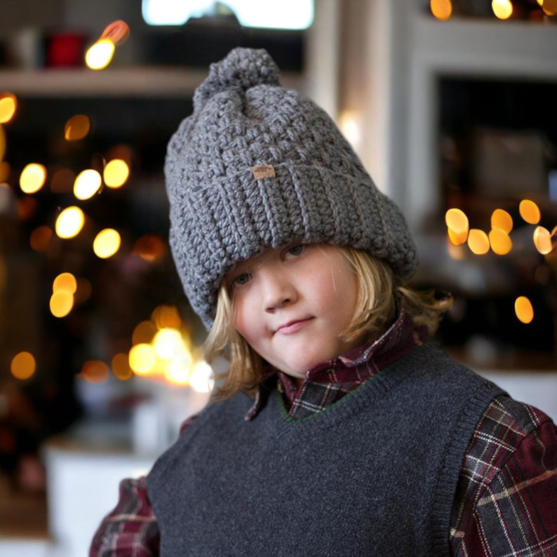 a child wearing a grey thick crochet hat with a rolled brim and knit look texture with pompom