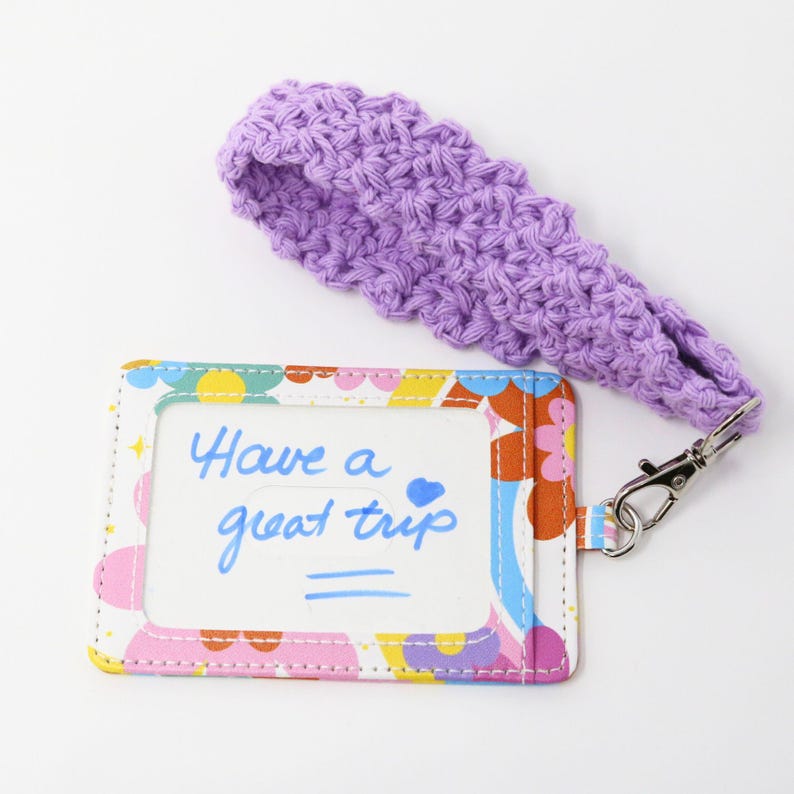 May include: A colorful ID holder with a floral pattern and the text "Have a great trip" in blue. It has a clear window and a lavender crocheted lanyard with a silver clasp.