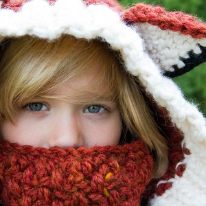 May include: A young person wearing a white and red crocheted fox hat with a matching red scarf. The hat has a white and black crocheted fox ear.