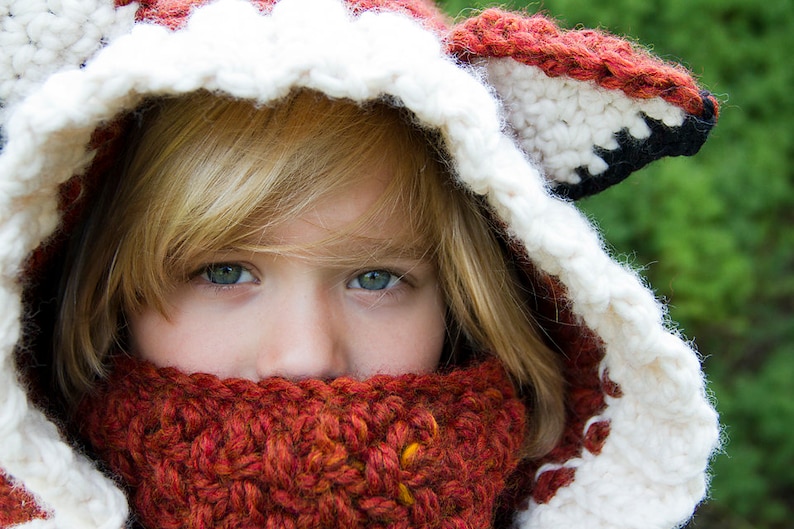 May include: A young person wearing a white and red crocheted fox hat with a matching red scarf. The hat has a white and black crocheted fox ear.