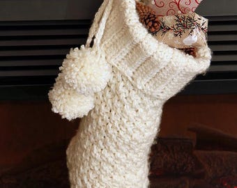 May include: A cream-colored, chunky knit Christmas stocking featuring a ribbed cuff, two matching yarn pom-poms attached to the side, and a hanging loop. The stocking is filled with a small pinecone and a gift wrapped in patterned paper.