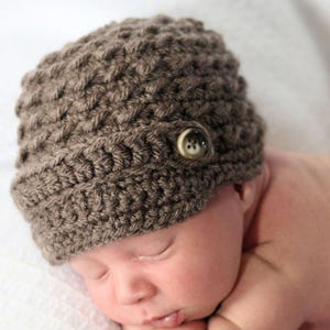 May include: A brown crocheted baby hat with a button on top. The hat is made with a textured stitch and has a slightly slouchy fit.