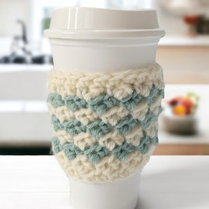 Puede incluir: Una taza de café blanca con un acogedor de ganchillo en crema y azul claro. El acogedor tiene un patrón de chevron.