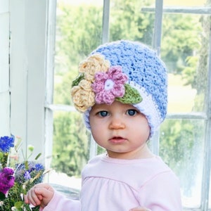 May include: A baby wearing a blue and white crocheted hat with a flower embellishment. The hat has a yellow, pink, and green flower design.