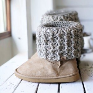 Op de afbeelding: Grijze gebreide laarzenmanchetten op een paar beige suède laarzen. De laarzen staan op een wit houten oppervlak.