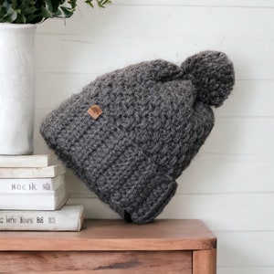 a grey thick crochet hat with a rolled brim and knit look texture with pompom on a pile of books