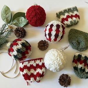 May include: A collection of handmade Christmas ornaments in various colors and patterns. The ornaments include red, white, green, and burgundy crochet balls and covers. Pinecones and greenery are also present, creating a festive holiday display.