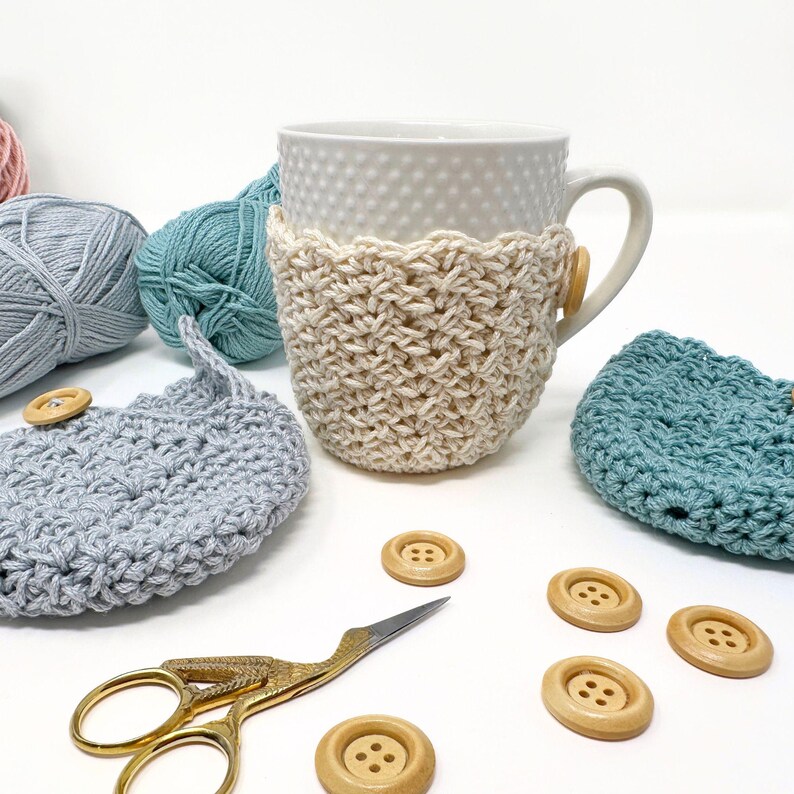 May include: A white ceramic mug with a cream-colored crocheted cozy and a wooden button. Additional crocheted items in blue and gray, along with wooden buttons and gold scissors, are scattered around the mug. Balls of yarn in various colors are also present.