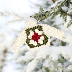 May include: White crocheted sweater ornament with a green and red flower design. The ornament is hanging from a snowy pine tree branch.