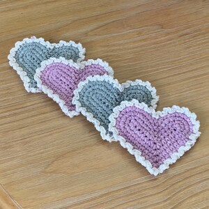 May include: Four handmade crochet heart-shaped coasters. Two are gray, and two are pink, each with a white scalloped edge. The coasters are arranged on a light brown wooden surface, showcasing a charming, handcrafted aesthetic.