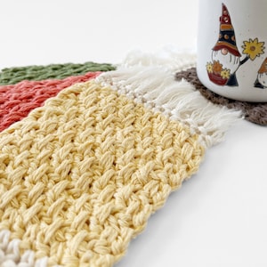 May include: Close-up of handmade crochet coasters in yellow, red, and green. The yellow coaster has a textured weave with white fringe. A white mug with gnome illustrations is partially visible, resting on a brown coaster.