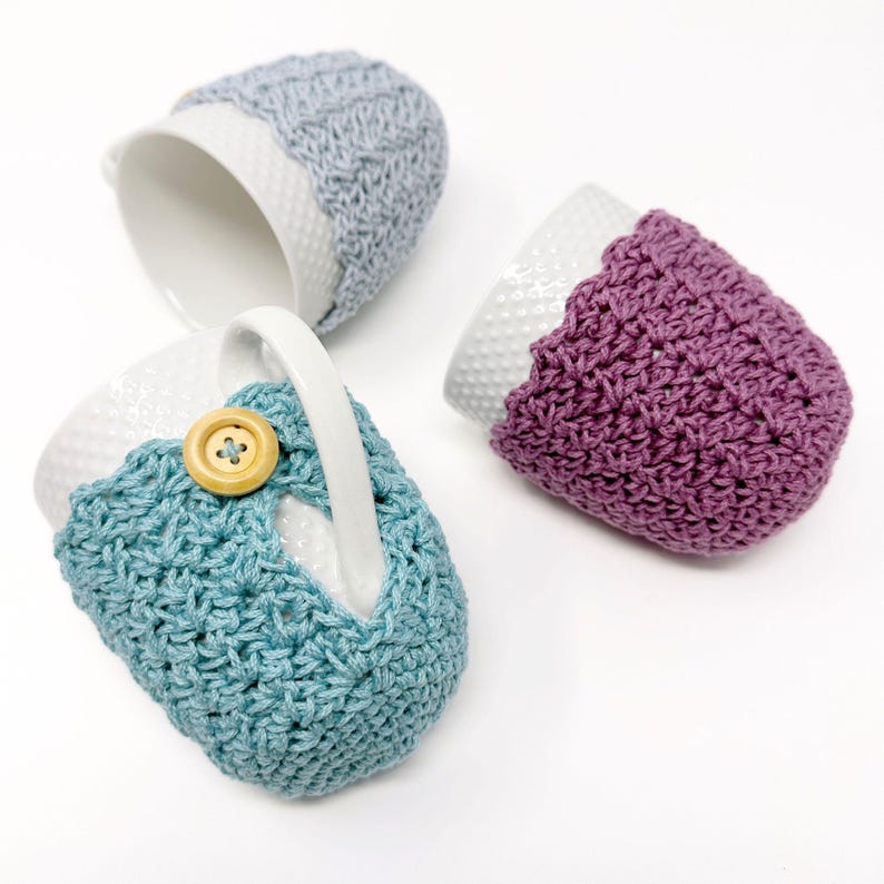 May include: Three white ceramic mugs with crocheted cozies in blue, purple, and gray. The blue cozy has a wooden button. The cozies are designed to fit around the mugs, adding a decorative and functional element.