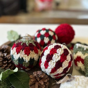 May include: A collection of handmade Christmas ornaments. The ornaments are crocheted in red, white, green, and burgundy patterns. Some are round, while others have a more elongated shape. The ornaments are displayed on a wooden surface with pinecones and greenery.