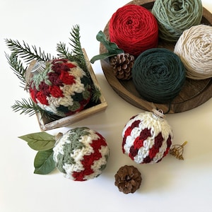 May include: A collection of handmade Christmas ornaments and yarn. The ornaments are crocheted in red, green, and white patterns. Several balls of yarn in red, green, and cream colors are in a wooden tray. Pinecones and greenery are also present.