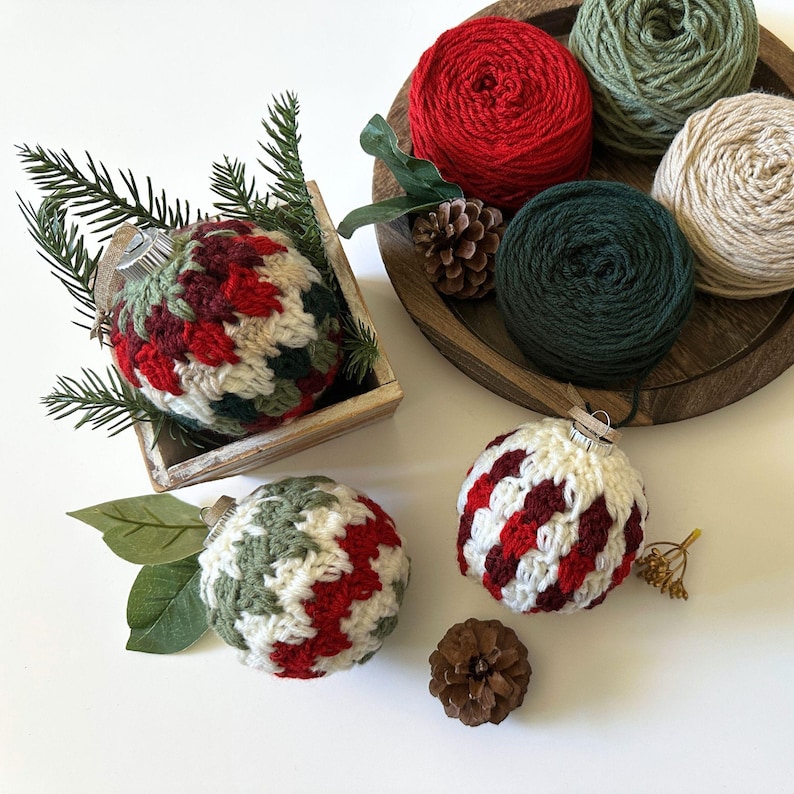 May include: A collection of handmade Christmas ornaments and yarn. The ornaments are crocheted in red, green, and white patterns. Several balls of yarn in red, green, and cream colors are in a wooden tray. Pinecones and greenery are also present.