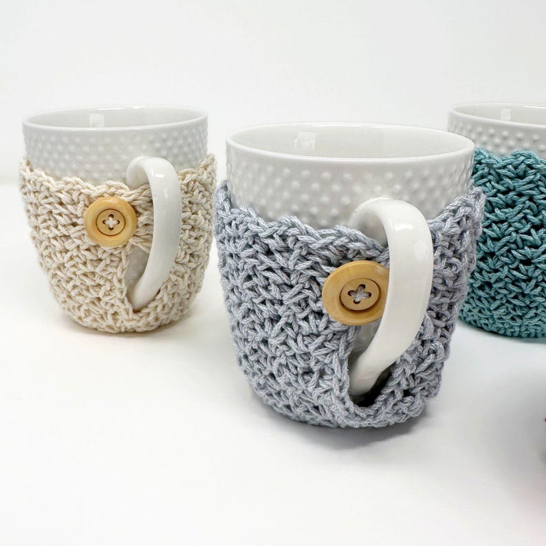 May include: White ceramic mugs with textured surfaces are adorned with crocheted cozies in cream, gray, and teal. Each cozy features a wooden button closure, adding a rustic touch to the drinkware.