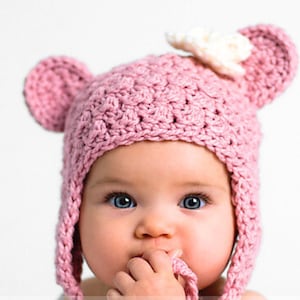 Puede incluir: Un gorro de bebé de crochet rosa con orejas de oso y una flor de crochet blanca en la parte superior. El gorro tiene una larga corbata que se puede atar debajo de la barbilla.