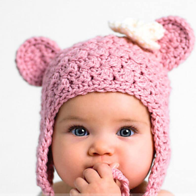 May include: A pink crocheted baby hat with bear ears and a white crocheted flower on top. The hat has a long tie that can be tied under the chin.