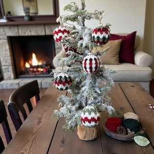 May include: A small, frosted Christmas tree decorated with handmade ornaments. The ornaments are red, white, and green, with a knitted or crocheted texture. The tree sits on a wooden table, with a cozy fireplace in the background.