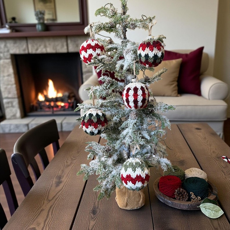 May include: A small, frosted Christmas tree decorated with handmade ornaments. The ornaments are red, white, and green, with a knitted or crocheted texture. The tree sits on a wooden table, with a cozy fireplace in the background.