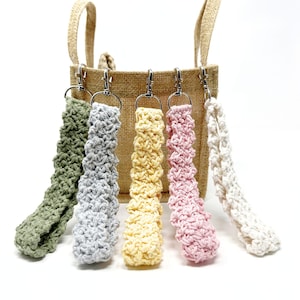 May include: Five handmade crochet wrist straps in various colors: green, gray, yellow, pink, and white. Each strap is attached to a silver-colored metal clasp. The straps are displayed in front of a tan burlap tote bag.