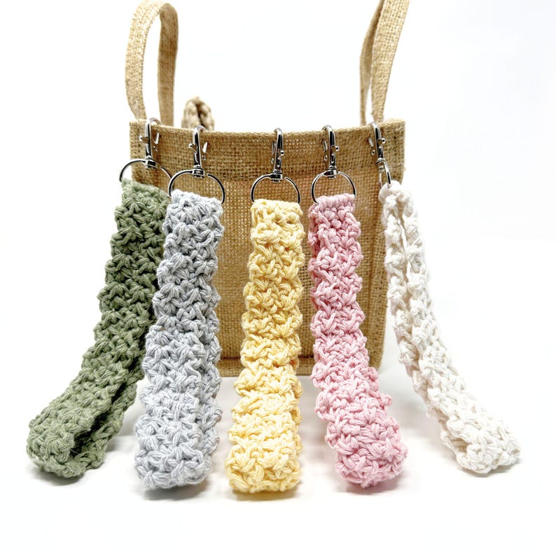 May include: Five handmade crochet wrist straps in various colors: green, gray, yellow, pink, and white. Each strap is attached to a silver-colored metal clasp. The straps are displayed in front of a tan burlap tote bag.
