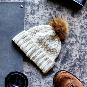 a cream thick crochet hat with a rolled brim and knit look texture with pompom snow in the background