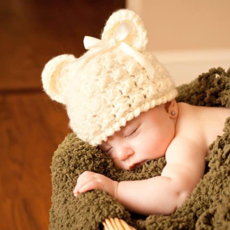 May include: A sleeping baby wearing a white crocheted bear hat with a white satin bow. The baby is lying on a brown fuzzy blanket.