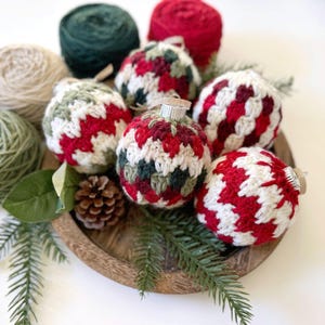May include: A wooden tray holds several crocheted Christmas ornaments in red, white, and green. The ornaments are round with silver metal caps. Green and beige yarn balls are in the background, along with pine needles and a pine cone.