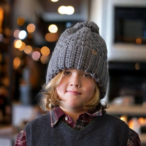 a child wearing a grey thick crochet hat with a rolled brim and knit look texture with pompom