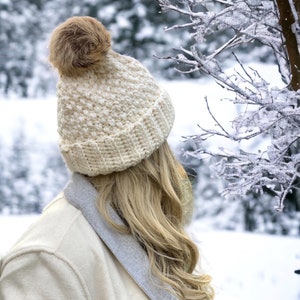 a women wearing a cream thick crochet hat with a rolled brim and knit look texture with pompom snow in the background