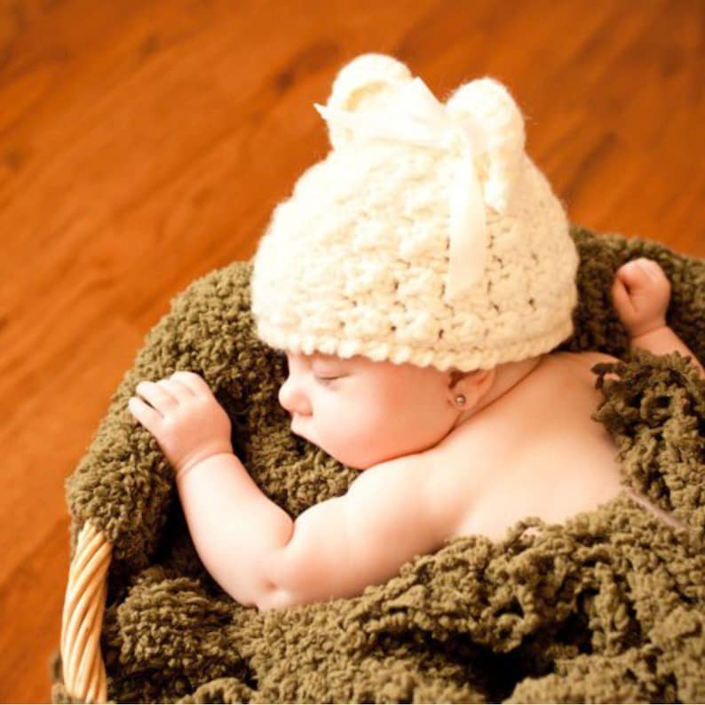 May include: A sleeping baby wearing a white crocheted hat with a white satin bow. The baby is lying on a green fuzzy blanket.