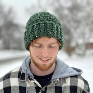 May include: A forest green crochet beanie with a thick ribbed cuff and subtle white flecks throughout the yarn. The hat features a textured stitch pattern and is constructed from a thick, warm-looking yarn, providing a snug fit around the head.