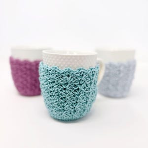 May include: Three white ceramic mugs with crocheted cozies in teal, purple, and gray. The cozies have a textured stitch pattern and a scalloped edge. The mugs are on a white background.