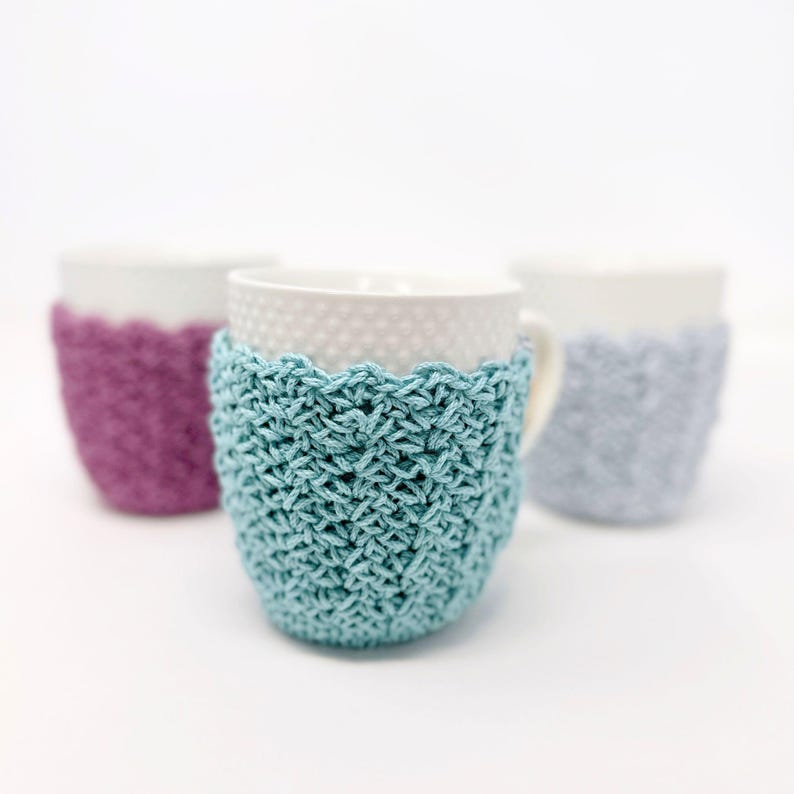 May include: Three white ceramic mugs with crocheted cozies in teal, purple, and gray. The cozies have a textured stitch pattern and a scalloped edge. The mugs are on a white background.