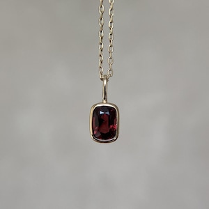 May include: A gold necklace featuring a rectangular pendant with rounded corners. The pendant contains a deep red, faceted gemstone set in a smooth gold bezel. The chain consists of small, uniform oval links that connect to the top of the pendant.