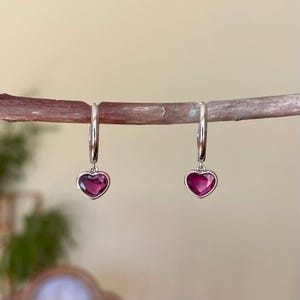 Puede incluir: Un par de pendientes de aro plateados, cada uno con un colgante de piedra preciosa rosa oscuro en forma de corazón. Los pendientes se exhiben en una rama de madera.