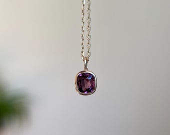 May include: A silver necklace featuring a square, deep purple gemstone pendant. The pendant is set in a gold-coloured bezel and hangs from a delicate chain. The gemstone has a cushion cut.