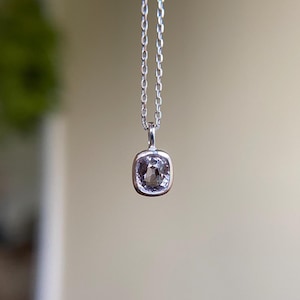 May include: A silver pendant necklace featuring a square, light-colored gemstone. The gemstone is set in a silver bezel and hangs from a delicate silver chain. The background is blurred, highlighting the necklace.